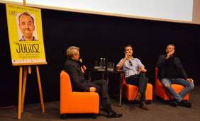 Pokaż zdjęcie: Trzy osoby siedzące na scenie z mikrofonami w ręku. W tle widać plakat filmu "Juliusz"