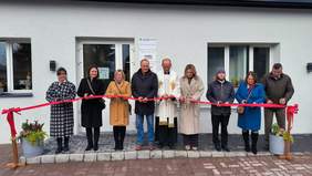 Przed budynkiem grupa 9 osób, 5 kobiet, 4 mężczyzn, stoją w szeregu, przecinają wstęgę, pora jesienna