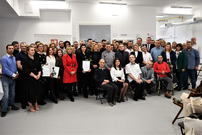 Pokaż zdjęcie: Na tle ściany stoi bardzo duża grupa osób pozujących do pamiątkowego zdjęcia