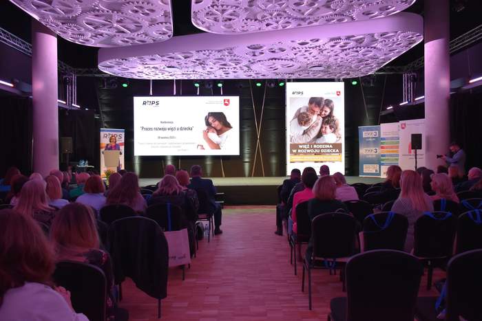 Pokaż zdjęcie: Zdjęcie sali konferencyjnej robione z tyłu sali, na zdjęciu widac krzesełka, siedzące osoby, oraz prezentacje na dużych ekranach, światło jest przygaszone