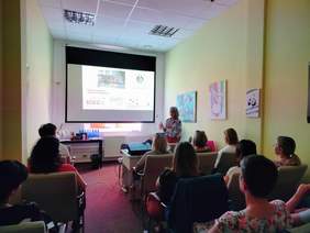 Pokaż zdjęcie: Przed zebranymi w sali stoi kobieta. Omawia prezentację multimedialną, która wyświetlona jest na rzutniku na ścianie. 