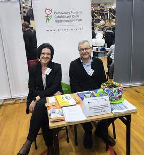 Pokaż zdjęcie: Przy stoisku wystawienniczym funduszu siedzą dwie osoby. Na stoliku leżą ulotki.