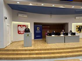 Pokaż zdjęcie: Za mównicą stoi elegancko ubrana kobieta w jasnych włosach za nią duże godło polski, obok długi stół za którym siedzi trójka ludzi. Przy stole rollup z napisem Samorząd Województwa Pomorskiego.