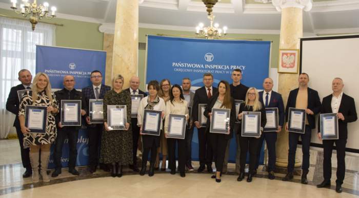 Pokaż zdjęcie: W sali kolumnowej zdjęcie laureatów stojących w półkolu, w rękach prezentują otrzymane dyplomy. W tle banery Państwowej Inspekcji Pracy 