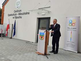 Pokaż zdjęcie: Mężczyzna w garniturze przemawia przy mównicy ustawionej przed budynkiem Centrum Opiekuńczo-Mieszkalnego w Olpuchu. Obok stoją flagi Polski i Unii Europejskiej oraz baner.
