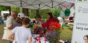 Pokaż zdjęcie: Dwie kobiety stoją pod namiotem z logo PFRON i zadają pytania z zagadnień poruszających tematykę niepełnosprawności dzieciom, które przyszły na piknik wraz z rodzicami. Na stole widoczne są cztery wazony z bukietami polnych kwiatów oraz kosze wiklinowe z ulotkami i gadżetami z logo PFRON.