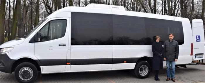 Pokaż zdjęcie: Na parkingu stoi biały autobus z logo PFRON oraz z naklejką informującą, że pojazd dostosowany jest do przewozu osób z niepełnosprawnością. W tle widoczne są drzewa pozbawione liści. Przed autobusem stoją kobieta wraz z mężczyzną.