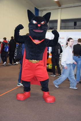 Pokaż zdjęcie: W sali stoi człowiek przebrany za dużego czarnego kota - maskotkę wydarzenia. Kot pręży mięśnie i pokazuje bicepsy. Jest ubrany w czerwone spodenki i czerwone buty. 
