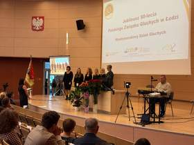 Na scenie stoi sześć kobiet ubranych na czarno. Jeden mężczyzna gra na instrumencie. Widać także osoby siedzące na widowni. 