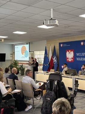 Pokaż zdjęcie: Uczestnicy siedzą na krzesłach i wózku, zwróceni w stronę osoby prowadzącej. Na ekranie wyświetlana jest prezentacja, a w tle widoczne są flagi Polski, Unii Europejskiej i NATO oraz baner „Wojewoda Warmińsko-Mazurski”.