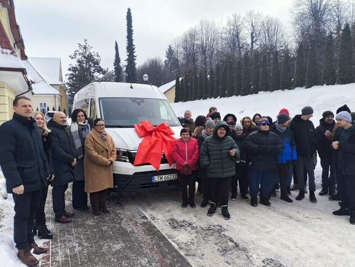 Pokaż zdjęcie: Grupa osób stojąca przed białym autobusem ozdobionym dużą czerwoną kokardą podczas zimowej uroczystości przekazania pojazdu. W tle ośnieżone drzewa i budynki.