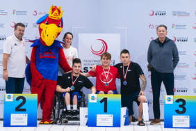 Pokaż zdjęcie: Zdjęcie grupowe po ceremonii medalowej, wykonane na tle z logo zawodów pływackich. Na zdjęciu stoi siedem osób: dwóch dorosłych (mężczyzna i kobieta) i pięciu młodych mężczyzn. Czterech młodych mężczyzn stoi w rzędzie, wszyscy ubrani w niebieskie koszulki i dresy, z medalami na szyjach. Młody mężczyzna pośrodku jest najwyższy. Obok nich, po prawej stronie, stoi uśmiechnięty mężczyzna w marynarce i maskotka – duża, czerwono-niebiesko-żółta papuga w koronie. Po lewej stronie grupy stoi mężczyzna opierający się na jednej lasce i kobieta. Zwycięzcy trzymają w rękach torby z logo sponsora.