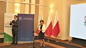 Pokaż zdjęcie: kobieta stoi na tle flag polskich i banera PFRONu i przemawia, obok tłumacz języka migowego