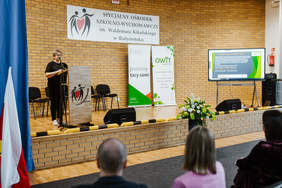 Pokaż zdjęcie: Po prawej Kobieta ubrana na czarno przemawia przez mikrofon stojąc przed mównicą. Za nią wisi baner z napisem Specjalny Ośrodek Szklono-Wychowawczy im. Waldemara Kikolskiego w Białymstoku oraz stoją dwa banery z logo PFRON i OWiT. 