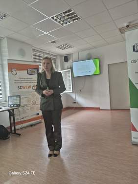 Pokaż zdjęcie: Kobieta w garniturze podczas prezentacji, w tle slajd z prezentacji.