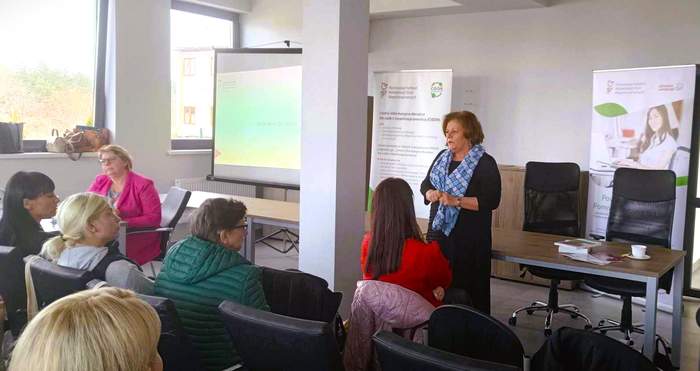 Na sali siedzą uczestnicy spotkania, a przed nimi stoi na środku Dyrektor Kujawsko-Pomorskiego Oddziału PFRON Aleksandra Gierej i przemawia. Za nia widoczne są banery, a po jej lewej stronie - w tle - stoi ekran z wyświetlonym slajdem.