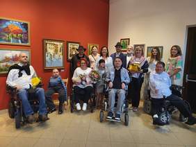 Pokaż zdjęcie: Liczna grupa artystów oraz uczestników wernisażu. Uczestnicy, w tym osoby bez rąk oraz na wózkach inwalidzkich stoją na tle obrazów.