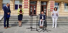 4 osoby na tle budynku urzędu, od lewej mężczyzna, kobieta, mężczyzna na wózku inwalidzkim mówi do mikrofonów, kobieta tłumacząca na j. migowy