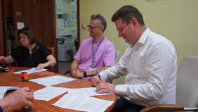 Pokaż zdjęcie: Przy owalnym stole widać siedzące trzy osoby: jedną panią i dwóch panów. Mężczyźni się uśmiechają. Pani sięga ręką po pieczątkę. 