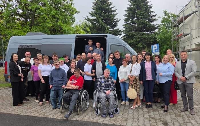 Pokaż zdjęcie: Duża grupa osób stoi na tle busa. Pośród tych osób osoby na wózkach inwalidzkich.