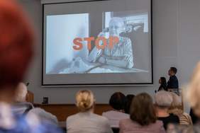 Na ekranie widoczny jest slajd: siedzi starsza osoba i napis STOP (chodzi o ochronę seniorów przed oszustwami, kradzieżami na wnuczka, itp.)