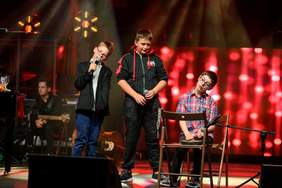 Pokaż zdjęcie: Występ artystyczny. Na scenie występuje trzech chłopców. Śpiewają i grają na instrumentach.