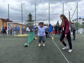 Pokaż zdjęcie: Kobieta ubrana w legginsy i białą koszulkę z napisem Monika jest w pozycji wyskoku w dal. Obok niej stoi kobieta ubrana w dres, która jest sędzią w dłoniach trzyma 2 kolorowe patyki. Za osobą skaczącą stoją kolejni zawodnicy. W tle widać bramk