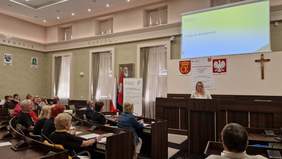 Pokaż zdjęcie: kobieta stoi na mównicy, przed nią słuchacze zasiadający na sali obrad w Urzędzie Miasta w Kielcach