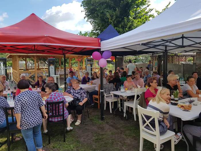 Pokaż zdjęcie: Duża grupa mężczyzn i kobiet siedzi na białych drewnianych krzesłach przy stolikach ustawionych pod parasolami ogrodowymi. W tle widoczne jest wysokie drzewo liściaste.