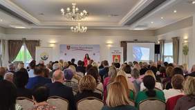 Pokaż zdjęcie: Grupa kilkudziesięciu osób siedzi w dużej sali konferencyjnej, na krzesłach ustawionych w kilku rzędach. Z przodu znajduje się kobieta z mikrofonem, a za nią baner gminy Stare Miasto oraz Gminnego Ośrodka Pomocy Społecznej.