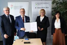 Pokaż zdjęcie: Dwóch mężczyzn i dwie kobiety. Przed sobą trzymają porozumienie zawarte pomiędzy PFRON, WUP w Krakowie i wszystkimi małopolskimi PUP. Za nimi rollupy  z logo PFRON i WUP