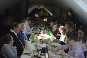 Przy podłużnych stołach siedzą ludzie. Trwa wielkanocne śniadanie. Na stołach leżą świąteczne potrawy, ciasta i dekoracje. 