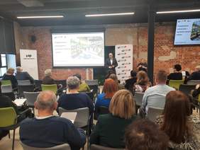 Pokaż zdjęcie: W sali konferencyjnej siedzą uczestnicy konferencji. Słuchają prelekcji. W tle widać ekran, na którym wyświetlona jest prezentacja. 