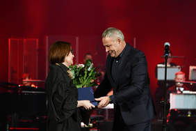 Kobieta i mężczyzna, ubrani na czarno, mężczyzna wręcza kobiecie niebieskie pudełko i róże