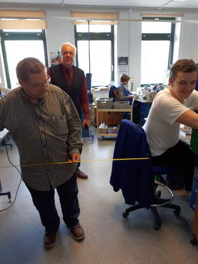 Pokaż zdjęcie: Wnętrze hali produkcyjnej,  pracownik siedzi przy stanowisku pracy, dwaj specjaliści BHP dokonują pomarów odległości