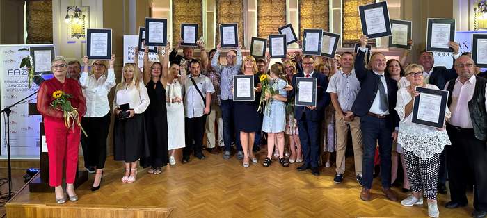 Pokaż zdjęcie: Na scenie, pod kopułą stoi duża grupa osób, część z nich trzyma uniesione w górę ramy z dyplomami, część statuetki. Wszyscy elegancko ubrani uśmiechają się.