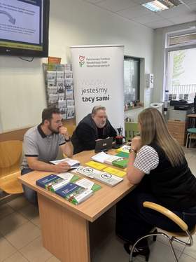 Dwóch pracowników PFRON prowadzi rozmowę informacyjną ze starszymi mieszkańcami przy biurku w pomieszczeniu urzędowym. Na stole widoczne są broszury i materiały dotyczące wsparcia osób z niepełnosprawnościami