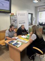 Dwóch pracowników PFRON prowadzi rozmowę informacyjną ze starszymi mieszkańcami przy biurku w pomieszczeniu urzędowym. Na stole widoczne są broszury i materiały dotyczące wsparcia osób z niepełnosprawnościami