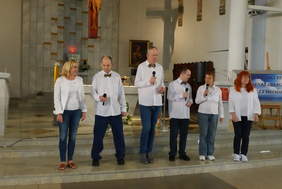 Pokaż zdjęcie: Sześć osób stoi na stopniach przed ołtarzem w kościele podczas występu. Wszyscy trzymają mikrofony i mają na sobie białe koszule – część z nich ozdobiona jest kolorowymi muszkami. Za nimi widać ołtarz, krzyż, obraz religijny oraz baner festiwalu.