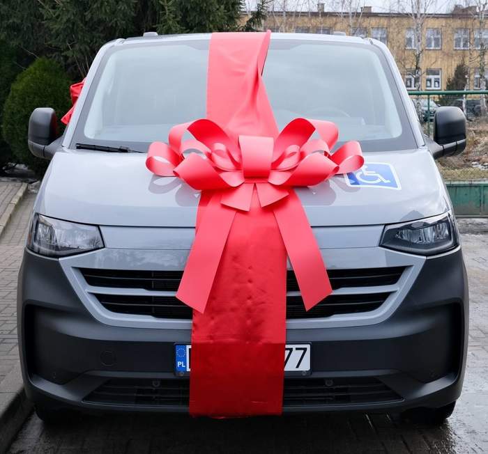 Pokaż zdjęcie: Srebrny bus widziany od przodu z dużą czerwono różową kokardą prezentową.   Na masce i przedniej szybie przebiega szeroka pionowa wstęga, a na środku maski znajduje się bardzo duża, ozdobna kokarda z wieloma pętlami i końcówkami zwisającymi w dół aż pod tablicę rejestracyjną.  Samochód koloru szarego ma nowoczesne, prostokątne reflektory i ciemnoszary zderzak.  Po prawej stronie na szybie, od strony pasażera, przyklejona jest niebieska naklejka z białym symbolem wózka.