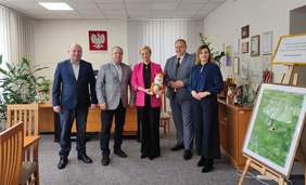 Pokaż zdjęcie: Grupa pięciu osób pozuje do zdjęcia w biurze. W tle widoczne są biurko, rośliny, liczne dyplomy i nagrody na półkach oraz godło Polski na ścianie. W centrum kobieta w różowej marynarce trzymająca maskotkę z logo Gminy Szumowo, obok niej stoją mężczyźni w garniturach i kobieta w granatowej bluzce. 