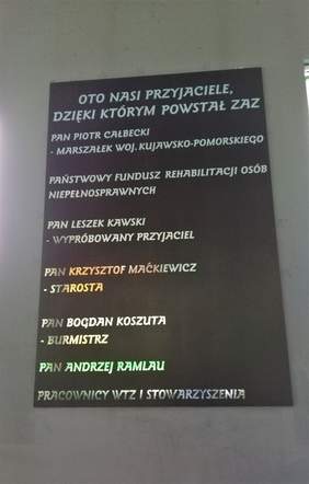 Pokaż zdjęcie: NA scianie wisi czarna tablica, a na niej widnieją nazwy i nazwiska osób, dzięki którym ZAZ powstał (nazwani przyjaciółmi), wśród wymienionych znajduje się PFRON