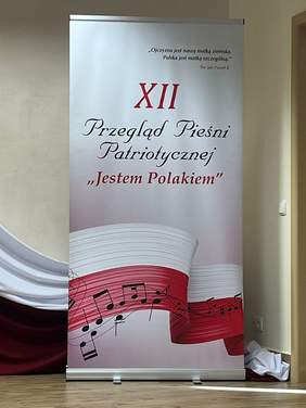 Pokaż zdjęcie: Na scenie WTZ Zgromadzenia Sióstr Służek NMPN w Szumowie stoi baner informacyjny. Na dole baneru namalowana jest wijąca się, biało – czerwona flaga, wzdłuż której przebiega pięciolinia i nuty. Na środku umieszczono napis „XII Przegląd Pieśni Patriotycznej „Jestem Polakiem.”” U góry znajduje się cytat św. Jana Pawła II „Ojczyzna jest naszą matką ziemską. Polska jest matką szczególną”.
