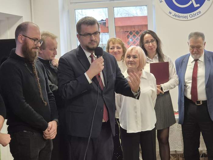 Pokaż zdjęcie: Dyrektor Oddziału Lubuskiego PFRON, ubrany w garnitur, przemawia z mikrofonem w ręku i gestykuluje, a za nim stoją inni uczestnicy spotkania, również ubrani odświętnie