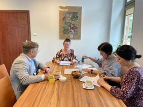 Pokaż zdjęcie:  Przy biurku siedzą trzy kobiety we wzorzystych bluzkach i mężczyzna w jasnej marynarce. Na biurku leżą pieczątki cukierki, kawa,  i woda mineralna. Za nimi na białej ścianie wisi obraz.