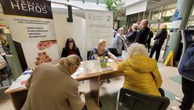 Pokaż zdjęcie: Przy stole na którym leżą katalogi i ulotki siedzą zainteresowane osoby i rozmawiają. W tle rollup z logo CIDON.