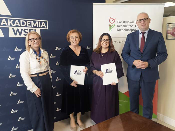 Pokaż zdjęcie: Cztery osoby stoją przed ściankami PFRON i Akademii WSEI; dwie osoby trzymają podpisane dokumenty. Spotkanie w pomieszczeniu biurowym, związane z zawarciem porozumienia o współpracy.