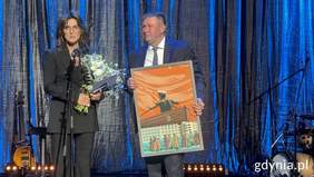 Pokaż zdjęcie: Kobieta ubrana w ciemną marynarkę i spodnie trzyma kwiaty i uśmiecha się. Przed nią stoi mikrofon. Obok stoi mężczyzna w garniturze pod krawatem trzyma obraz. Za nimi wisi kurtyna.