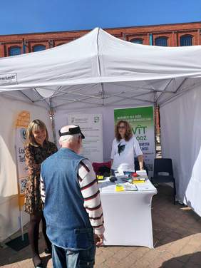 Pokaż zdjęcie: Przy stoisku PFRON stoją dwie kobiety i jeden starszy mężczyzna. Rozmawiają ze sobą i uśmiechają się. 