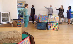 Pokaż zdjęcie: W dużym pomieszczeniu pomalowanym na biało 4 kobiety i 1 mężczyzna ogladaja prace plastyczne ustawione na podłodze i pod ścianami